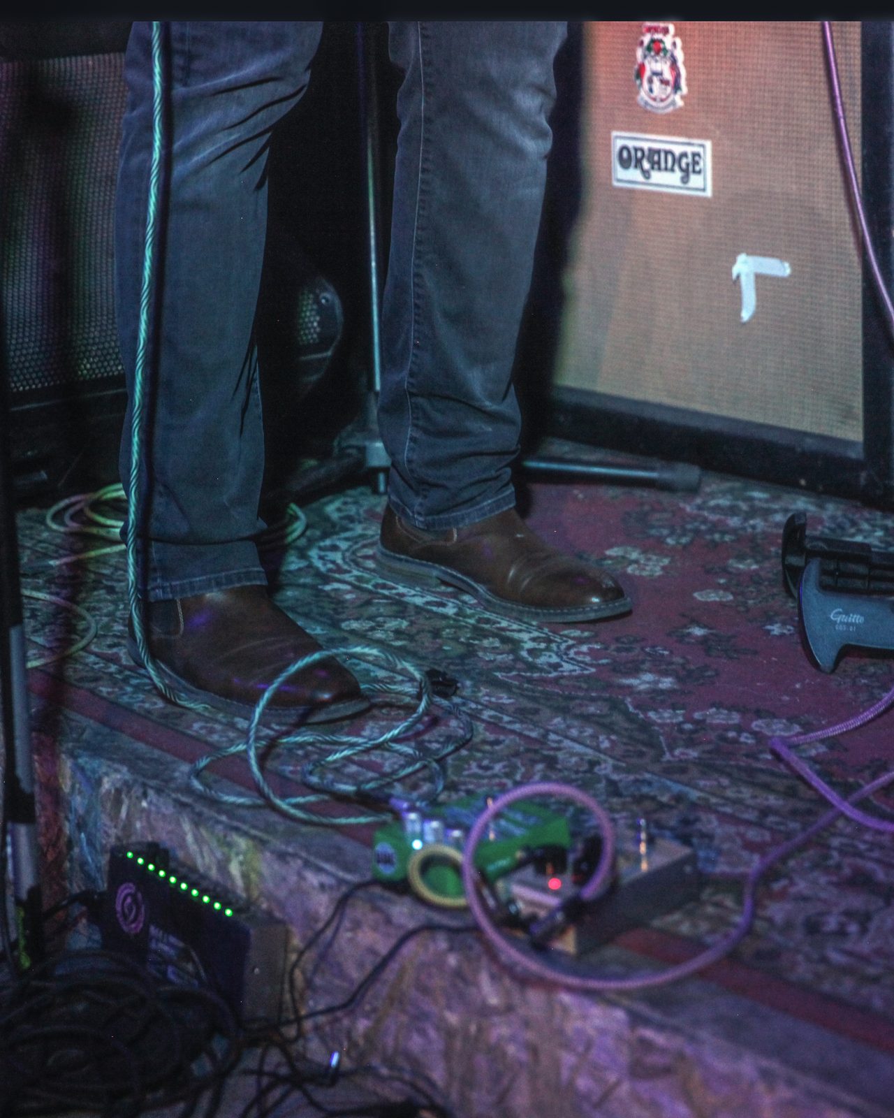 Boots on a patterned carpet in front of an Orange amplifier, pedalboard and cables running across the stage floor